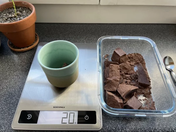 Ceremonial cacao chunks being weighed to 20 grams in a turquoise cup on a digital kitchen scale.