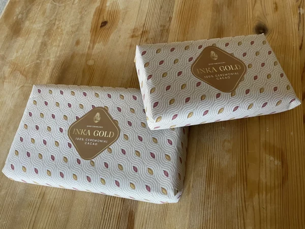 Two artisanal Inka Gold ceremonial cacao blocks wrapped in patterned paper, placed on a rustic wooden table.