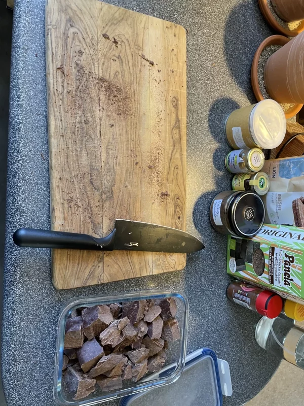 Freshly chopped ceremonial cacao on a wooden cutting board beside ingredients like panela, spices, and superfoods.