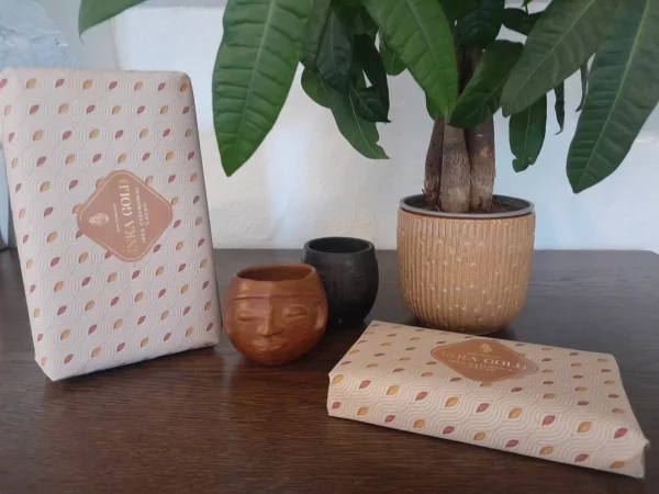 Two Inka Gold ceremonial cacao blocks (1kg and 500g) wrapped in patterned paper, styled with handcrafted cups and a potted plant on a wooden surface.